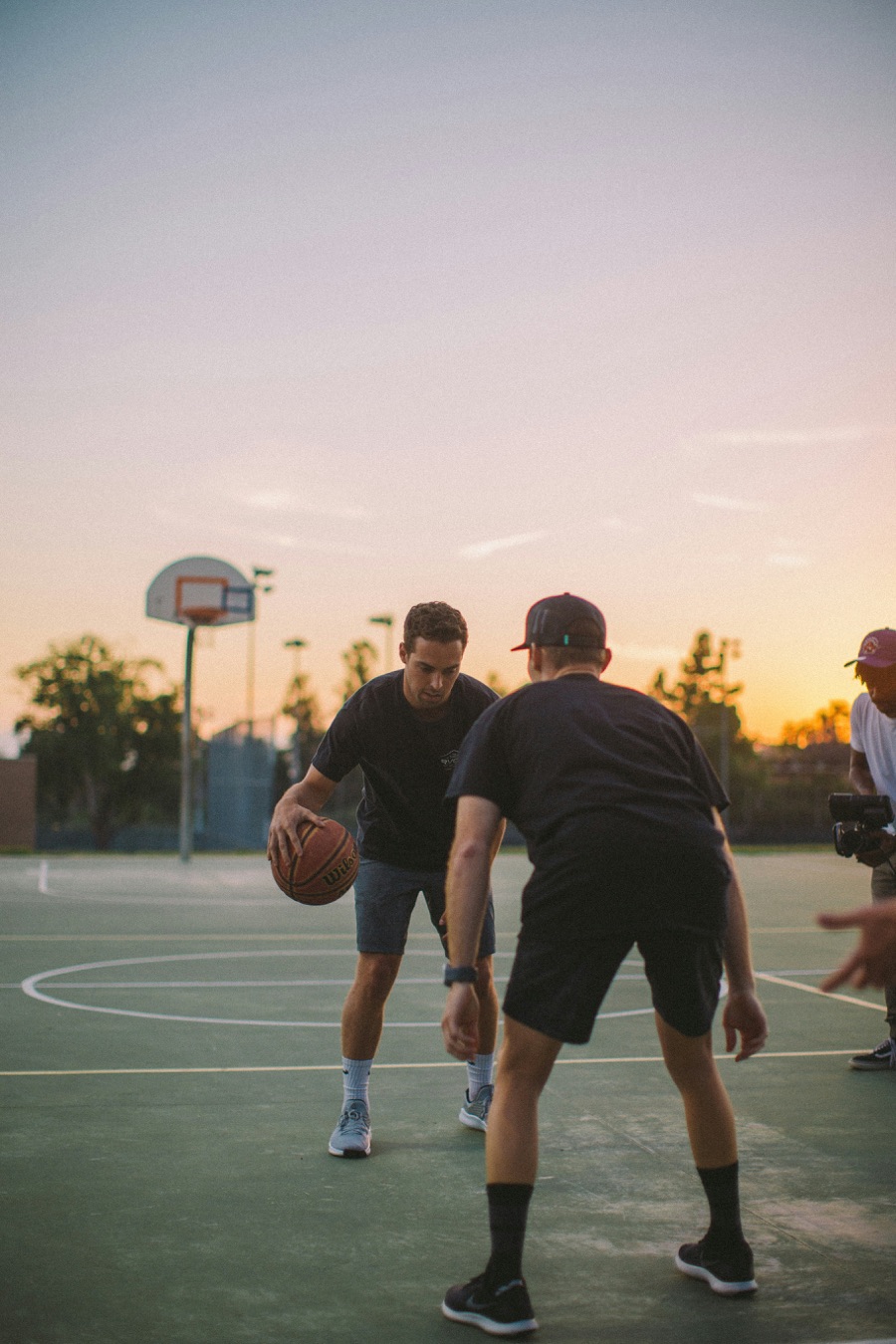 Adults playing basketball on an outdoor court at sunset.
